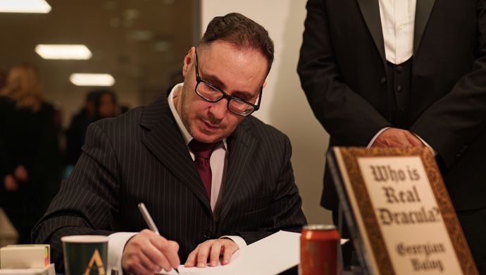 A man writing in a book with a sign in front saying who is real dracula?