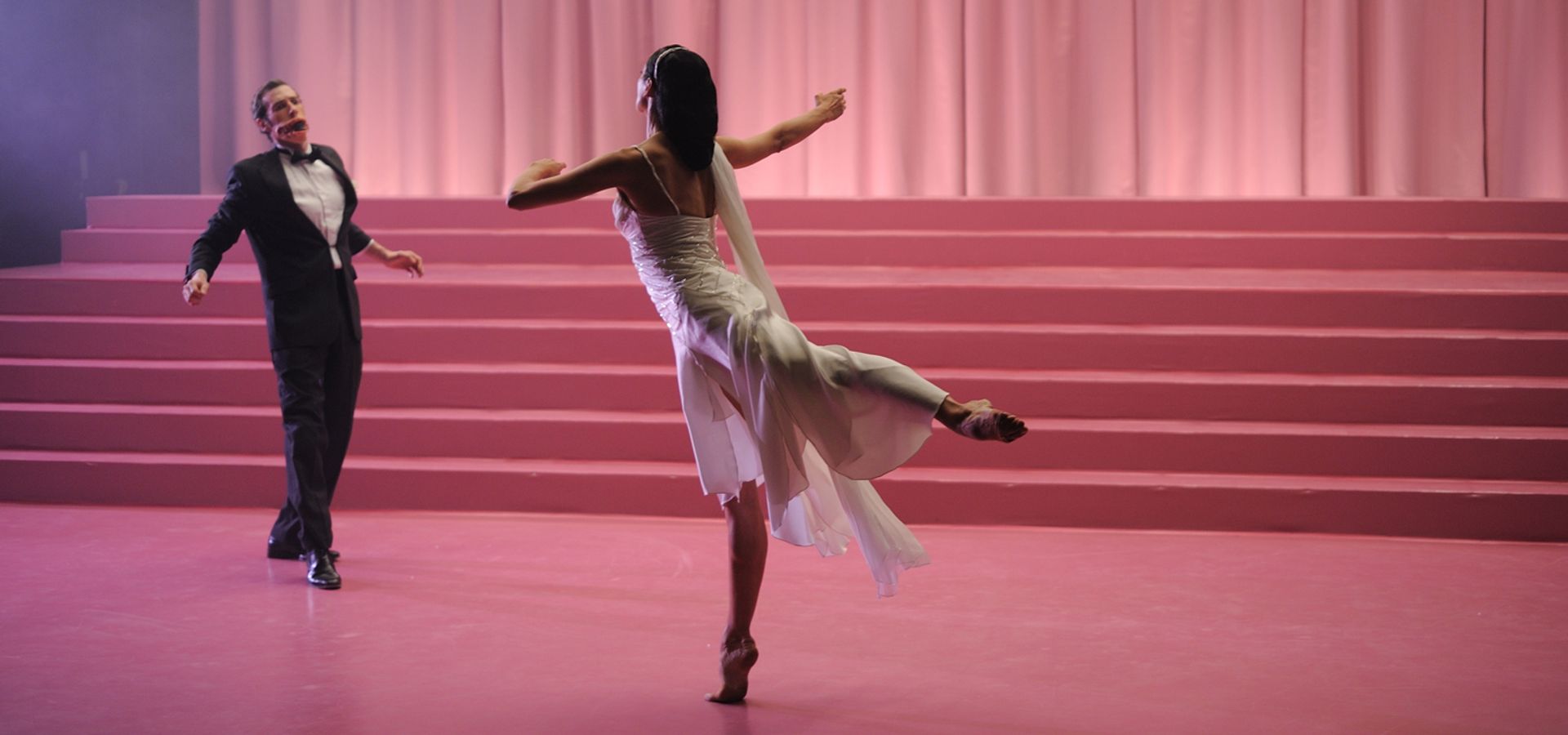 A Stuart Croft film image, a female ballerina lifting on leg in there air, looking at a male dancer in a suit with a disformed mouth.