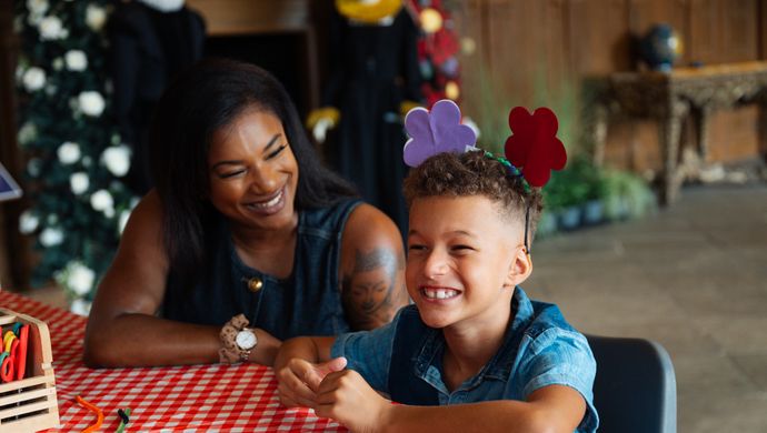 Adult and child sat at a crating table smiling. The child is wearing crafted flower headband.