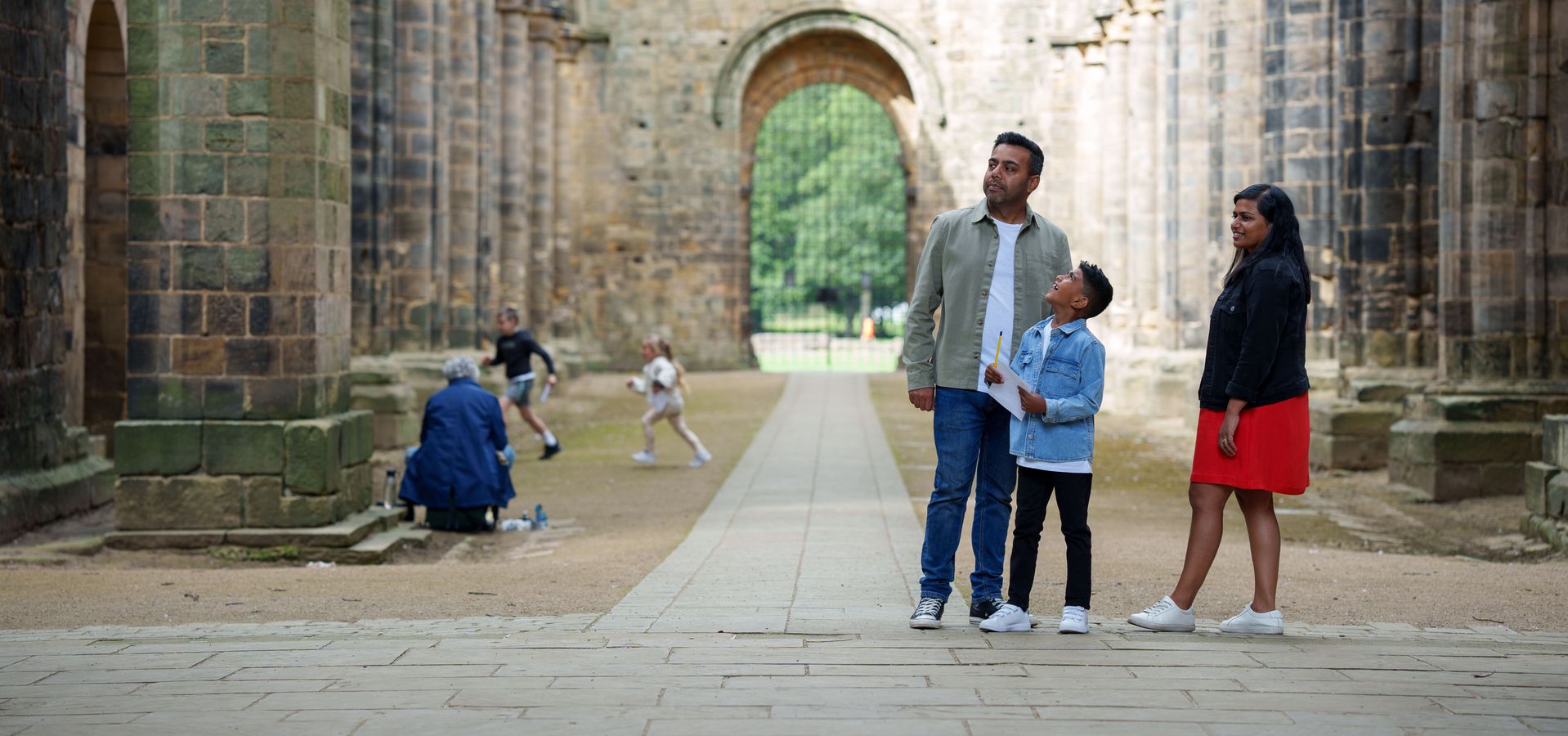 Two adults and a child looking around Kirkstall Abbey with children running in the background