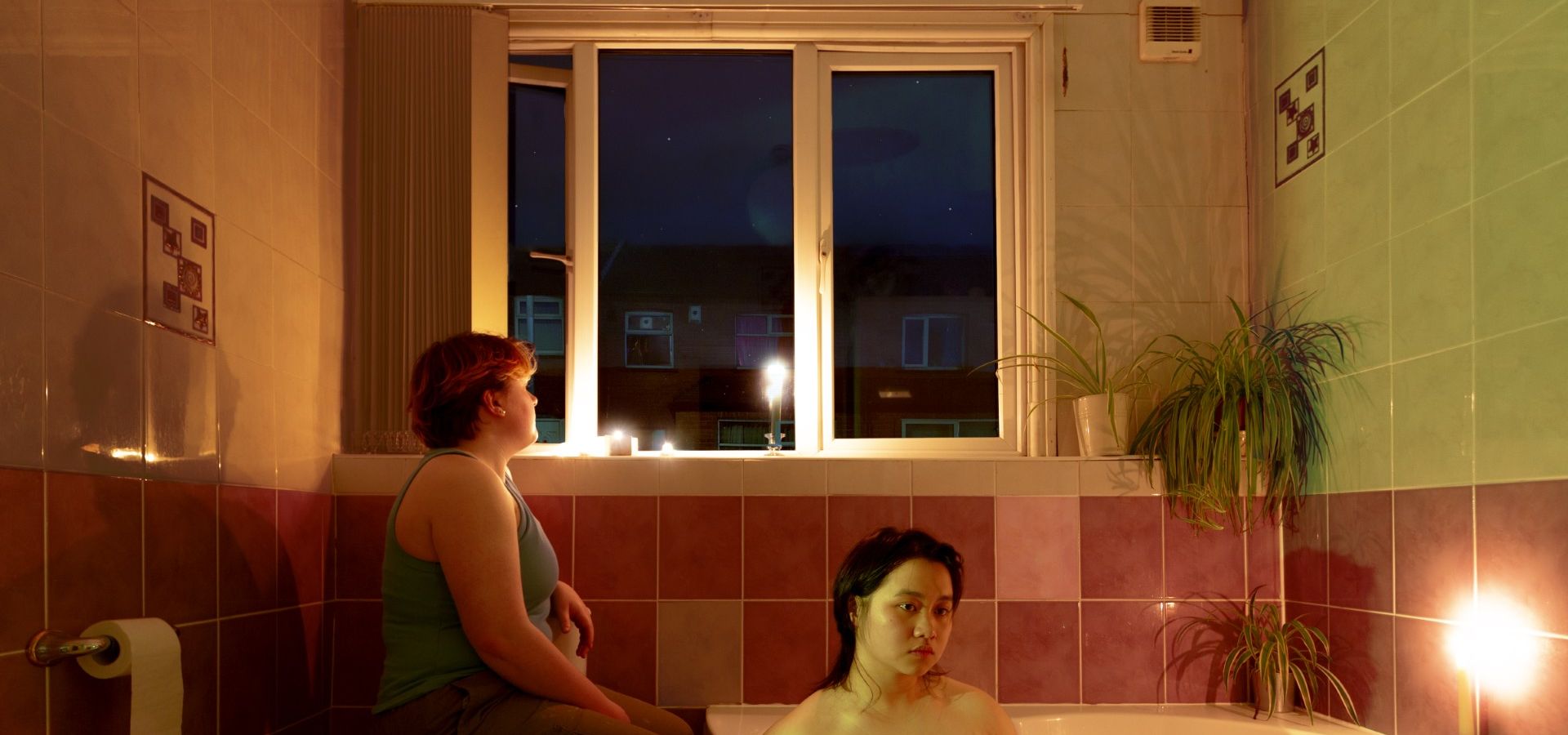 A woman sat in a corner bath, and a clothed woman sat on the toilet with the lid down, looking out a window into a dark sky with houses in view