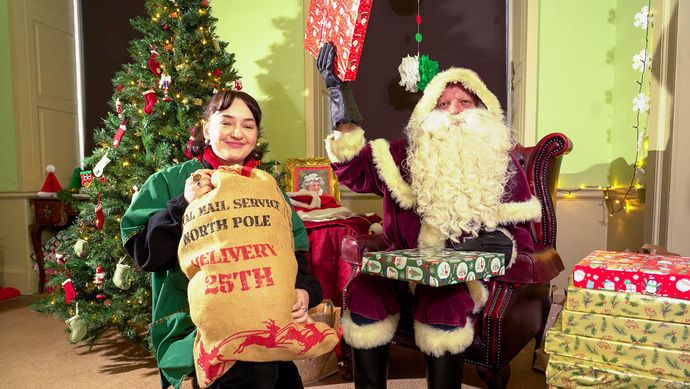 Father Christmas and an elf sat next to a christmas tree with a bag of presents