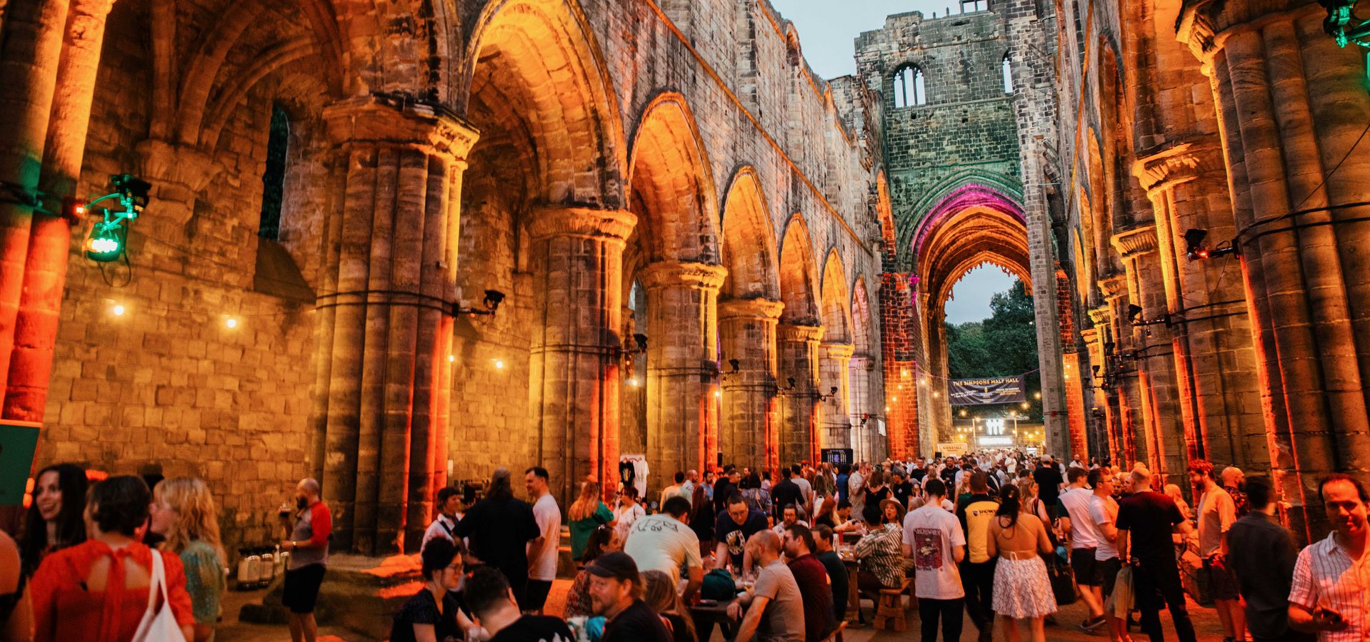A Cistercian monastery lit up orange at dusk with lots of people enjoying beer and food.