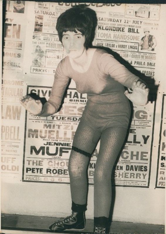 Sue Brittain in a very simple wrestling outfit in from of a wall with newspaper headings behind
