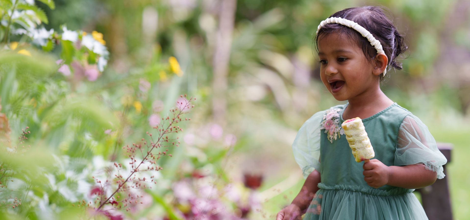 A toddler holding an ice cream looking at flowers in a garden