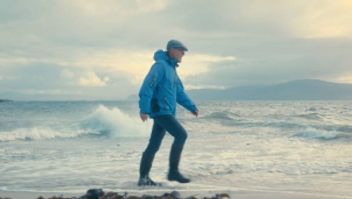 Photograph of male figure walking, with a background of a rough sea and cloud sky.