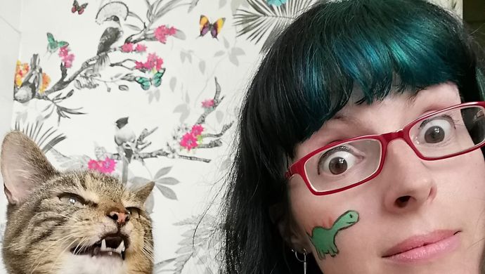 Nicky Bray, a woman with dark hair wearing red glasses, with face paint on, next to a cat