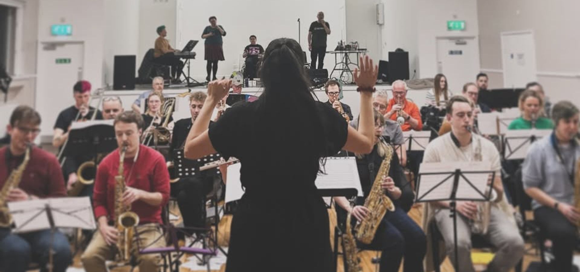 A conductor of Leeds Project Big Band stood in front of the band in a large, white room