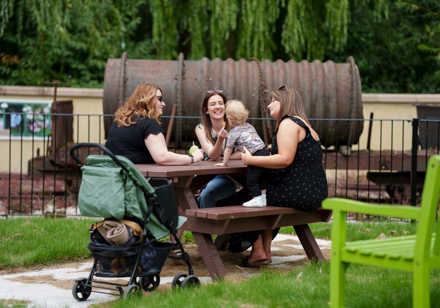3 women and a toddler on a picnic bench with a pushchair next to it, with industrial machinery behind