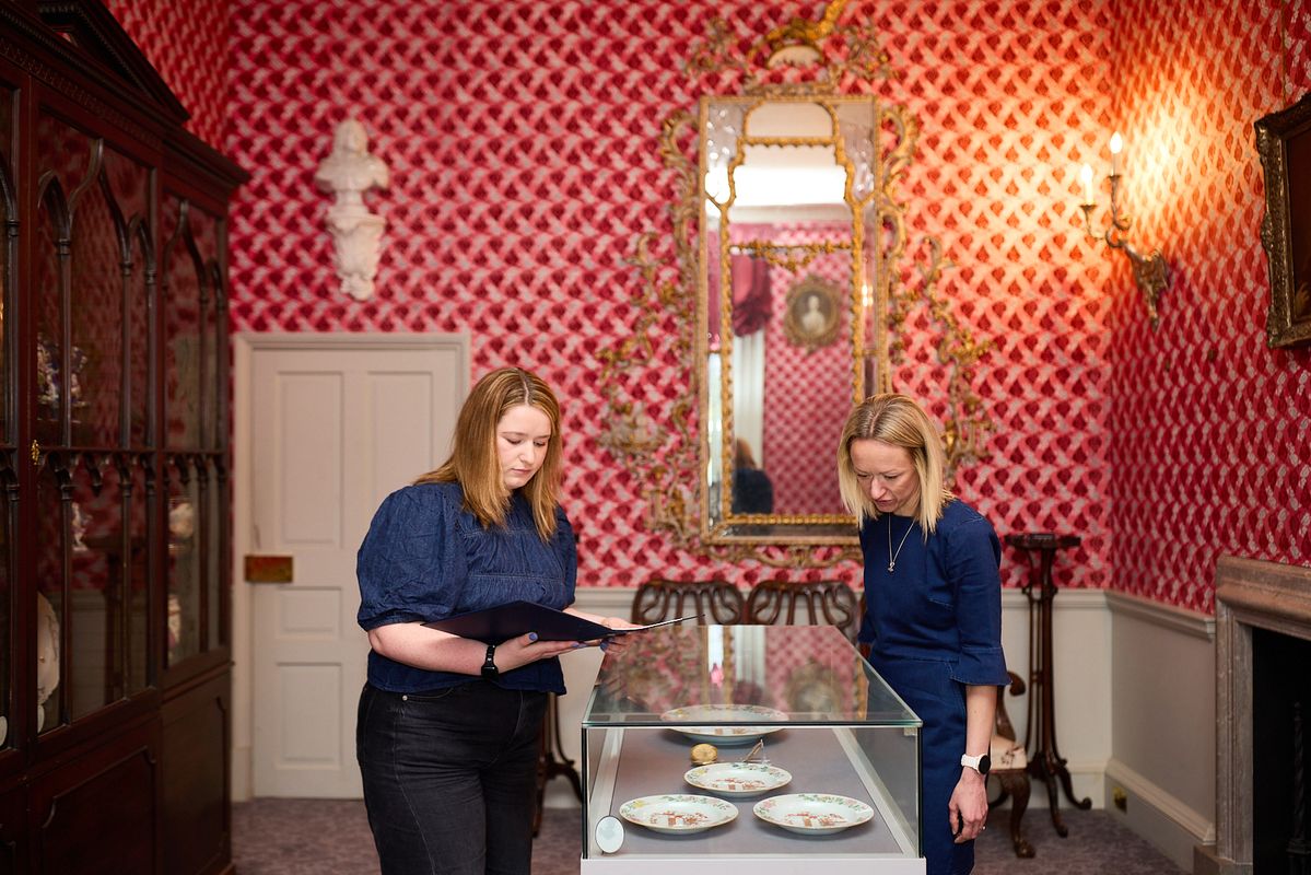 Two adults looking in a low display case in Temple Newsam, in a room with pink and red wall papered room