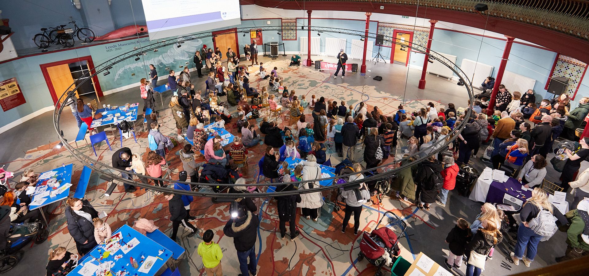 a large circular room from above with people stood watching a performance and craft tables in the back
