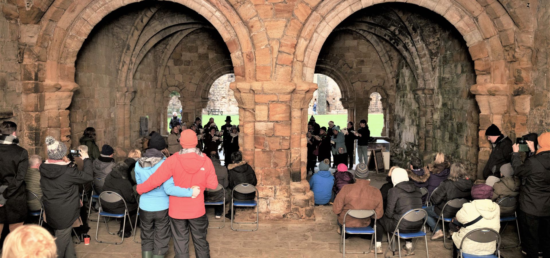 A group of seated and stood people listening to a choir under archways in Kirkstall Abeby ruins