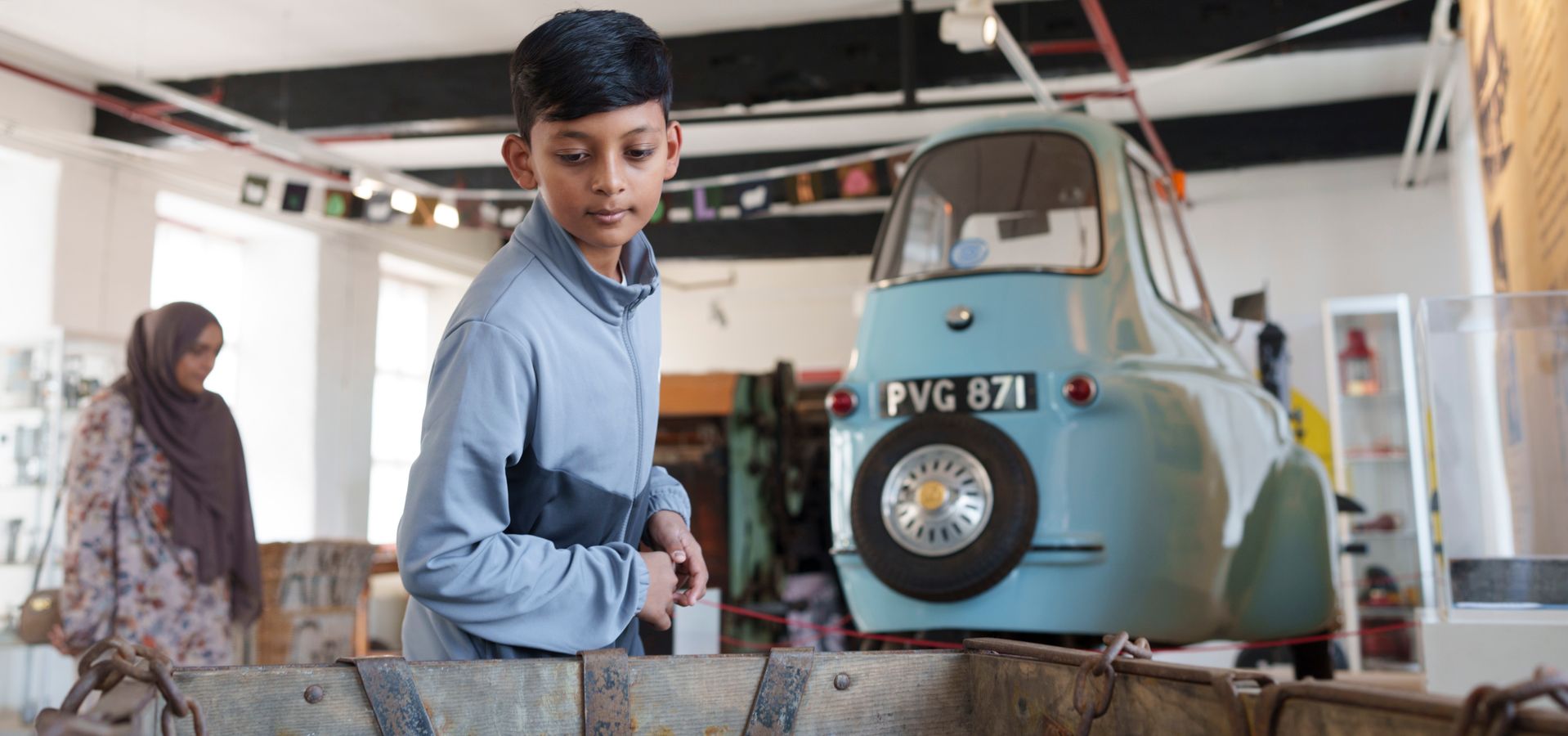 a teenager looking at an object in the museum with a small blue car behind on a platform