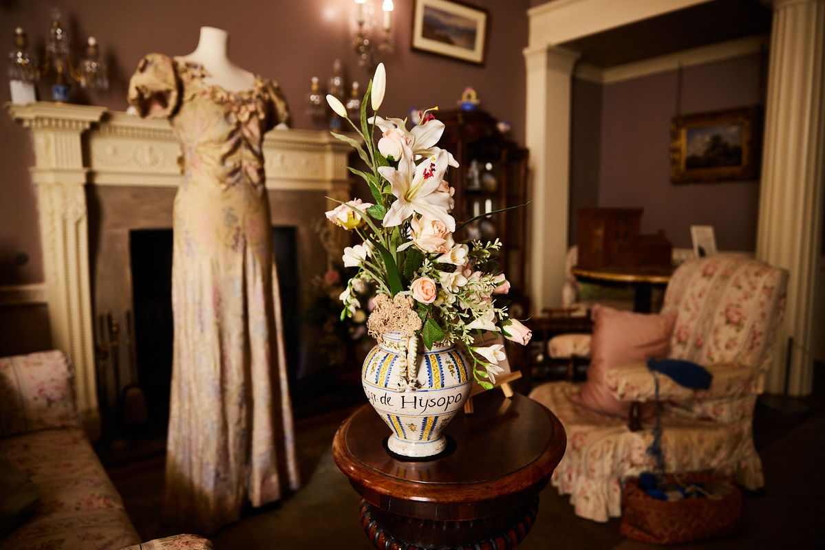 A vase with a floral arrangement with a mannequin with a dress on behind and a old fashioned chair