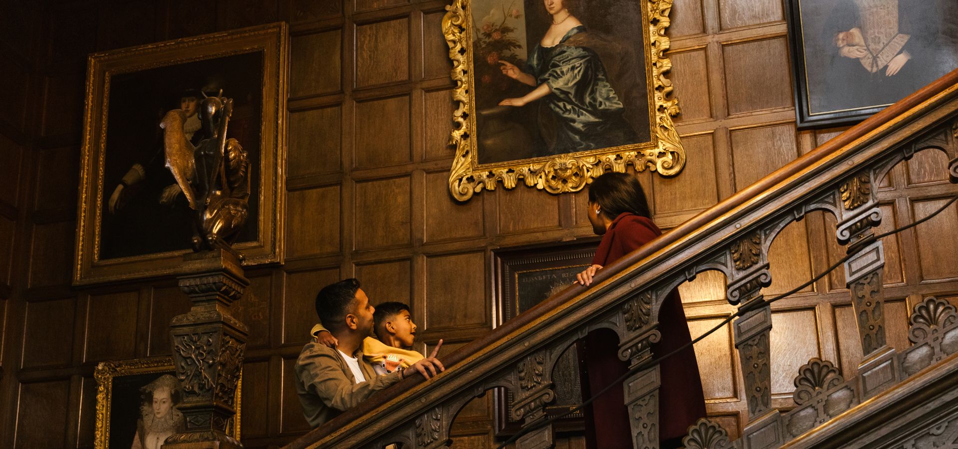 Two adults and a child looking at portriats on a stairway in Temple Newsam