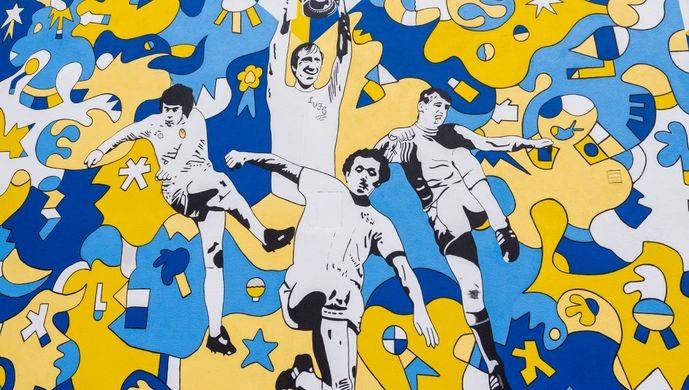 A mural on the side of a house in blue, yellow, black and white colours, showing people playing sport under rays of sun.