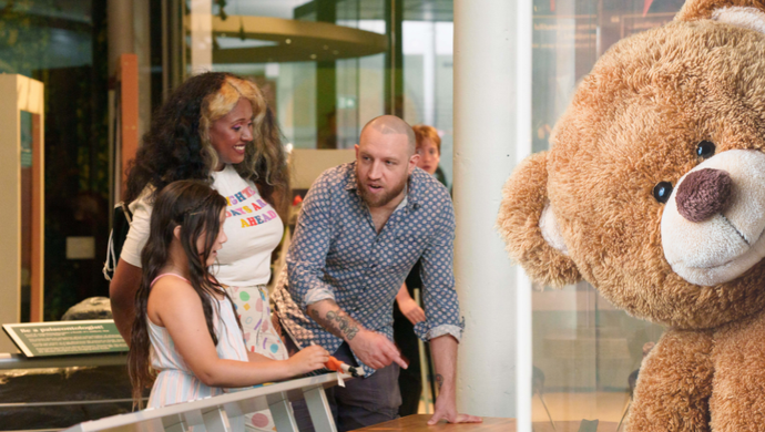 Two adults and a child looking at a museum display with a large teddy bear peaking in