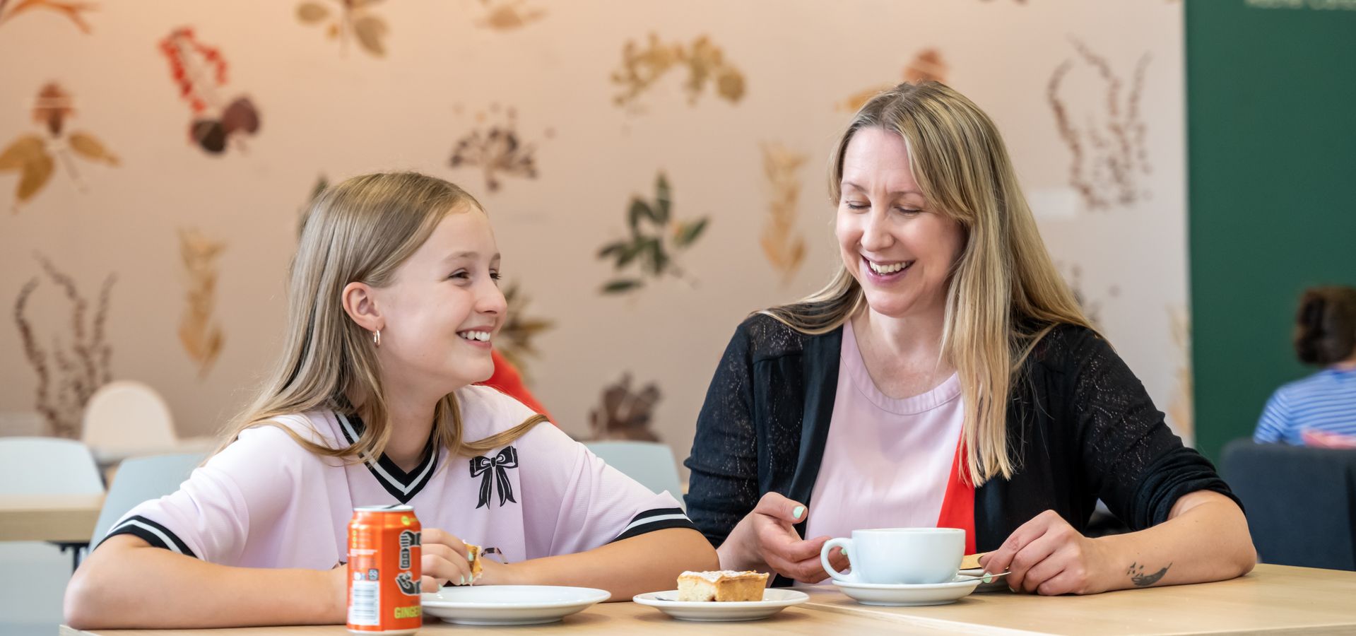 A woman and child looking at eachother sat next to eachother on a table, with pressed botcanicals on the wall behind
