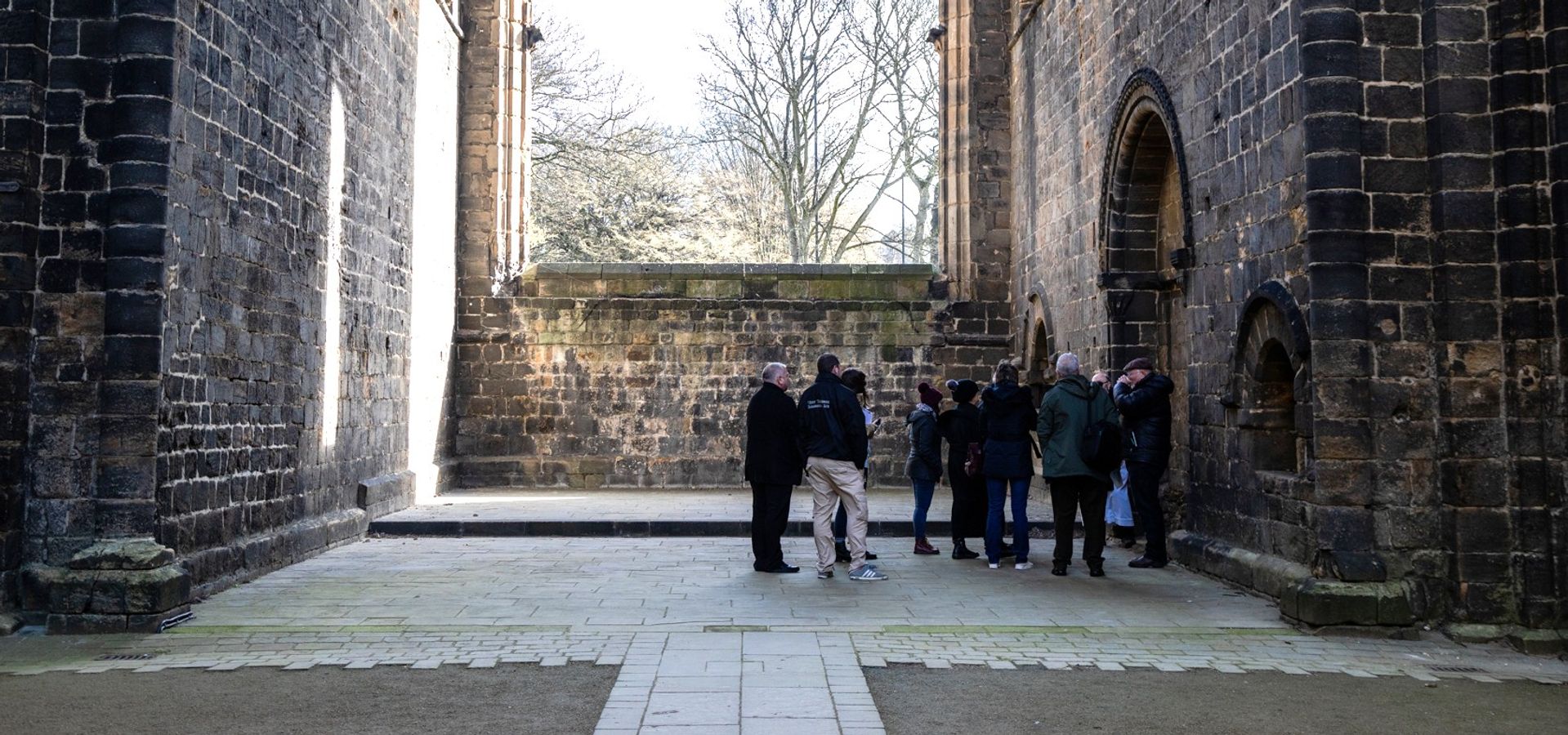 A group on a tour in Kirkstall Abbey, stood in front of a large window with no glass