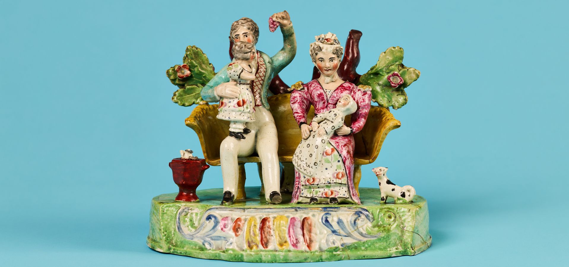 A ceramic sculpture of a couple and a child sat on a bench with leaves behind