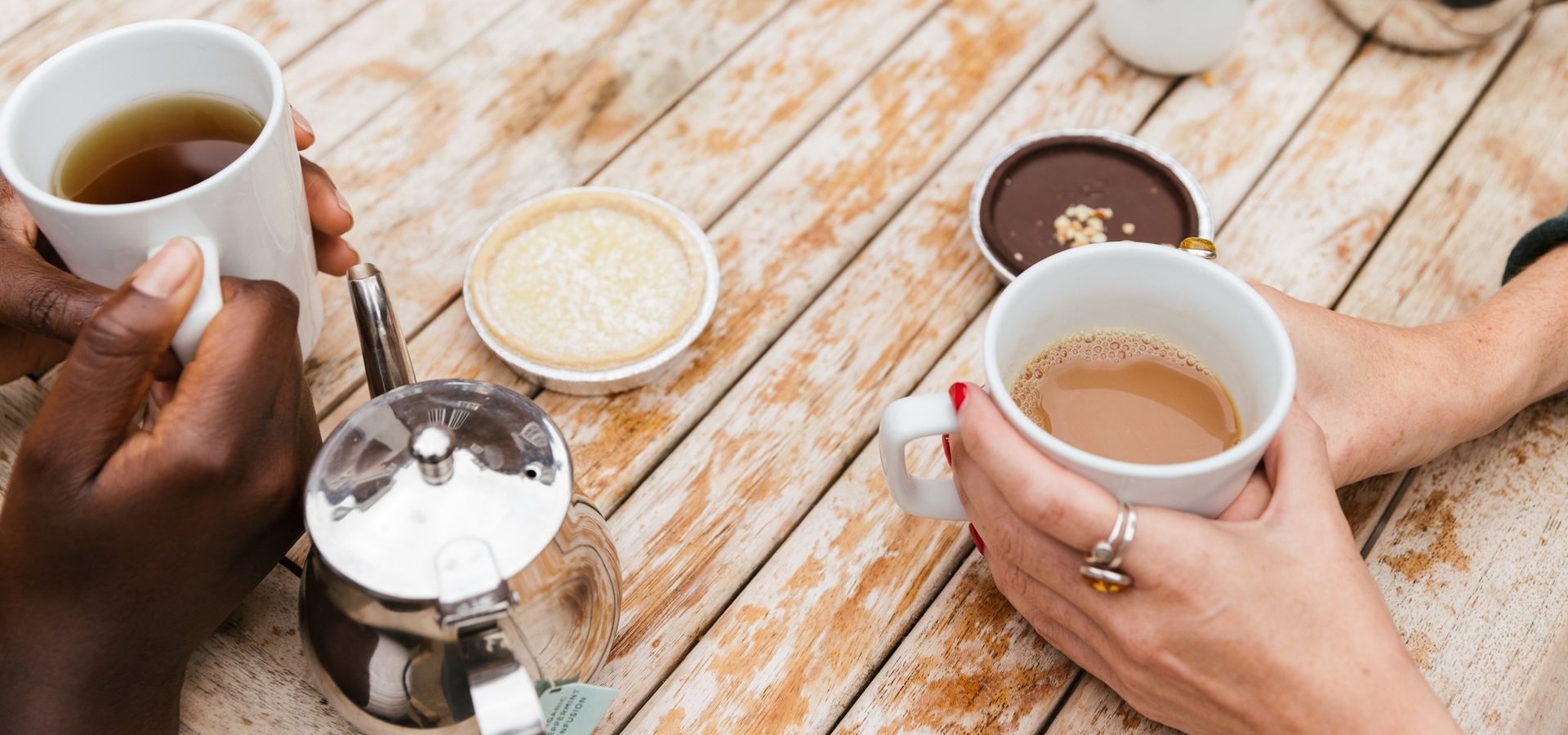 2 peoples hands holding cups of tea with two tarts