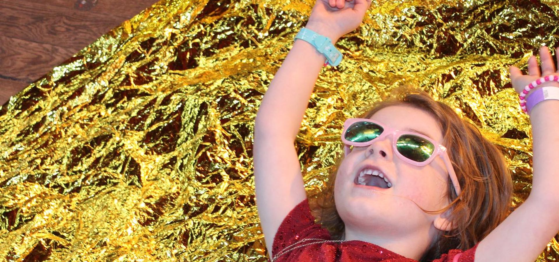 A toddler in sunglasses in a red sparkly dress laid on gold fabric