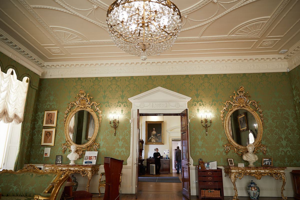A room with green wallpaper and a large white doorframe in the centre, with tables, mirrors and busts on each side