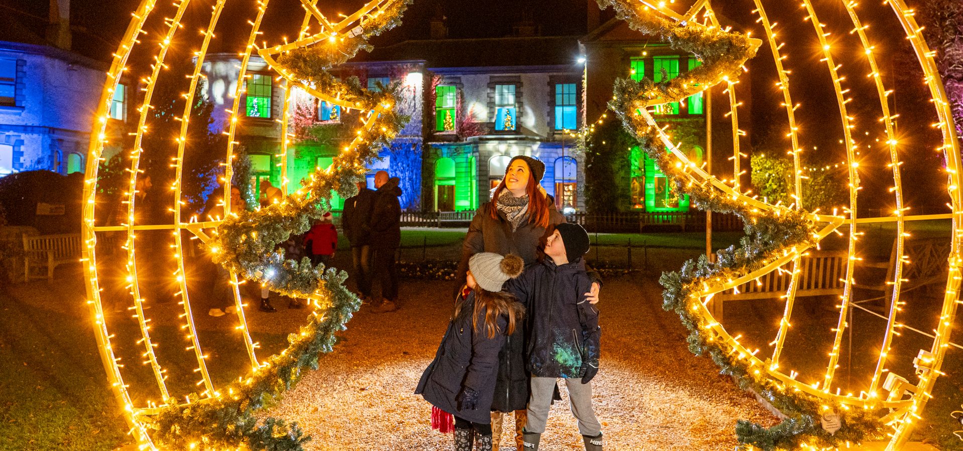 An adult and 2 children stood under a christmas tree light