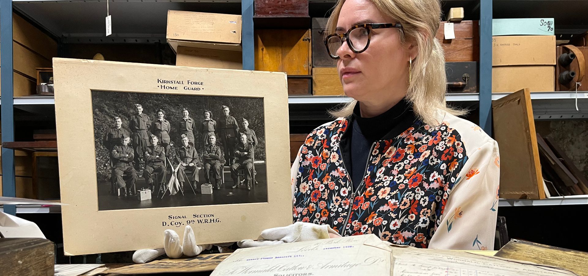 A curator holding a photograph of 12 people in 2 lines, with log books surrounding the curator