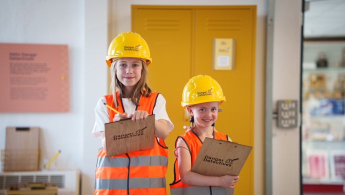 Two children wearing yellow hard hats and orange high-visibility jackets, holding clipboards that say danger detectives