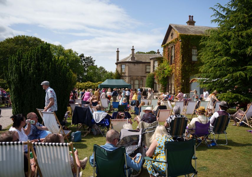 A crowd of people sat in deck chair on a lawn in front of a heritage house