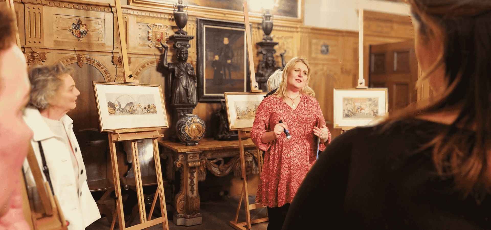 Looking over the shoulder of two people, a woman gives a talk surrounded by artwork on easels