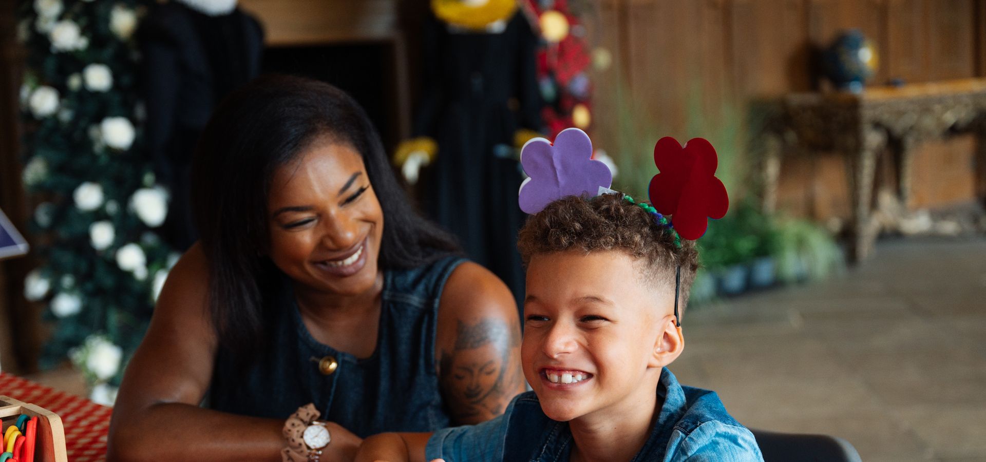 Adult and child sat at a crating table smiling. The child is wearing crafted flower headband.
