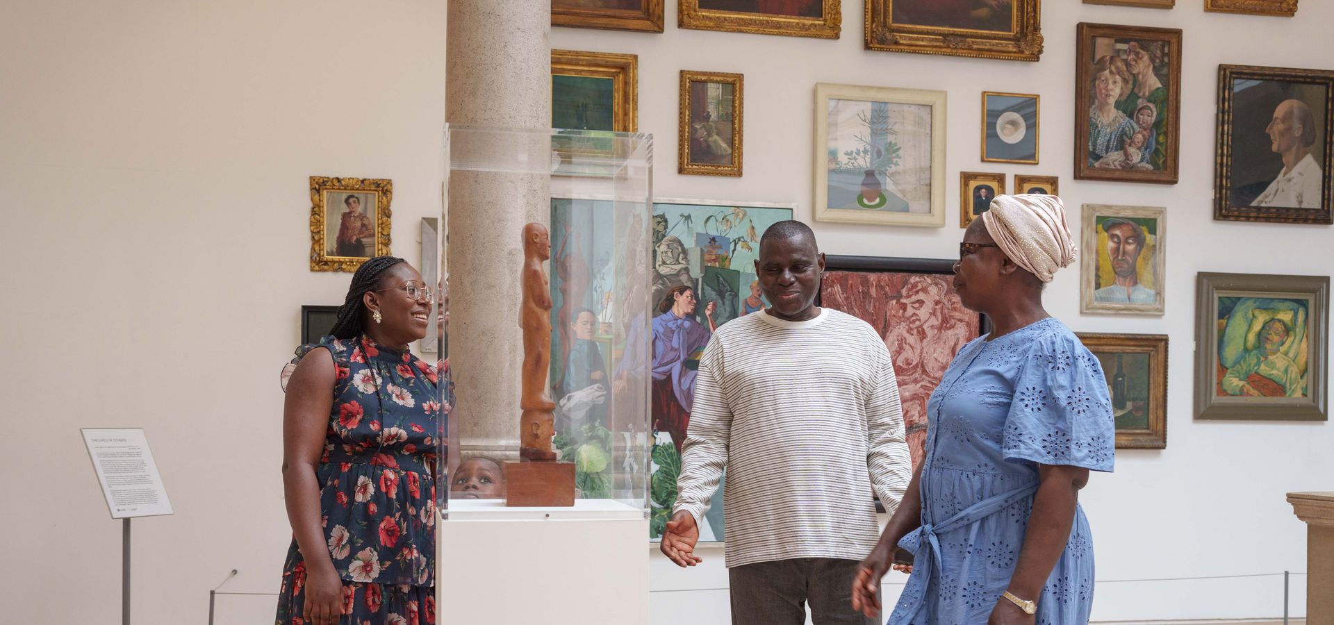 3 adults looking at a sculpture on a plinth with a wall of framed artworks behind