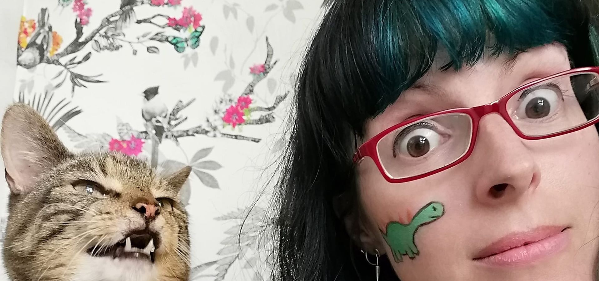 Nicky Bray, a woman with dark hair wearing red glasses, with face paint on, next to a cat