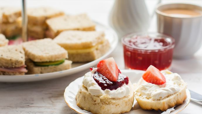 A scone with cream and jam and a plate of sandwiches behind