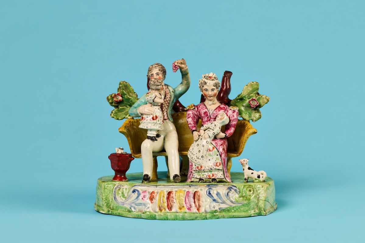 A ceramic sculpture of a couple and a child sat on a bench with leaves behind