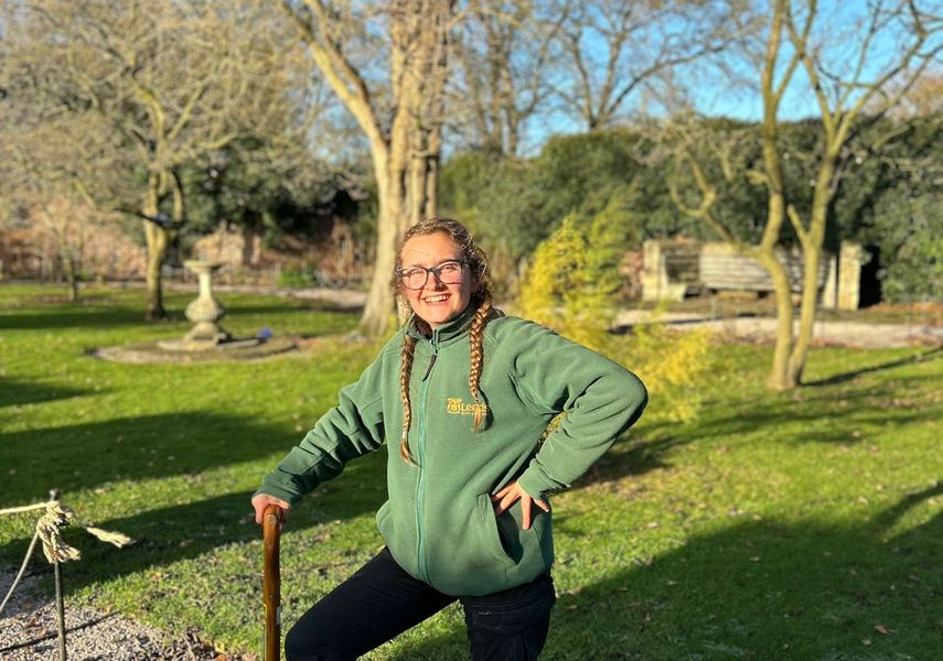 A woman with a green jumper on stood in a garden with her foot on a shovel