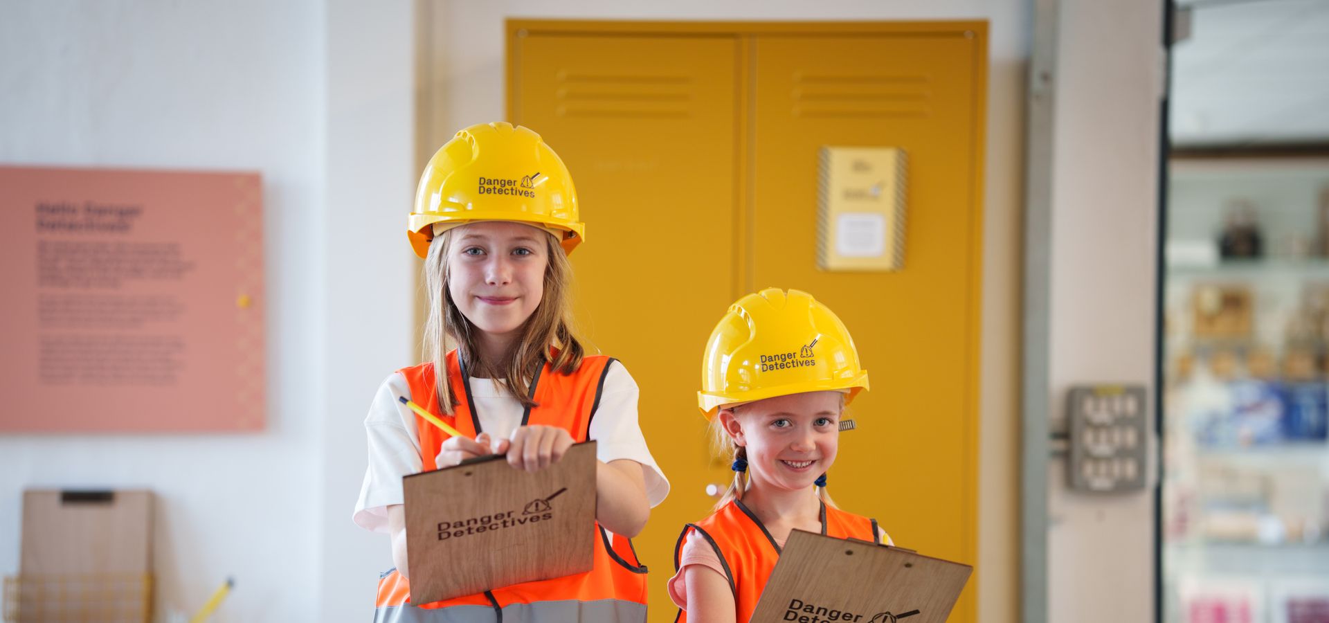 Two children wearing yellow hard hats and orange high-visibility jackets, holding clipboards that say danger detectives