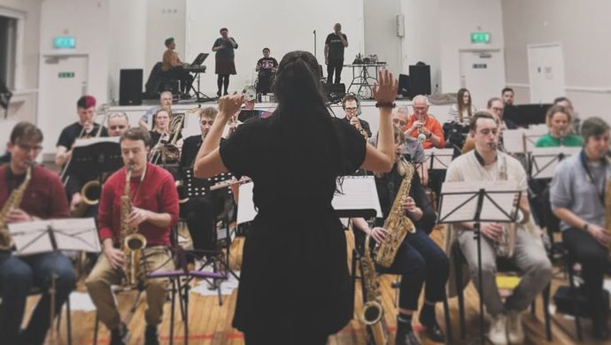 A conductor of Leeds Project Big Band stood in front of the band in a large, white room
