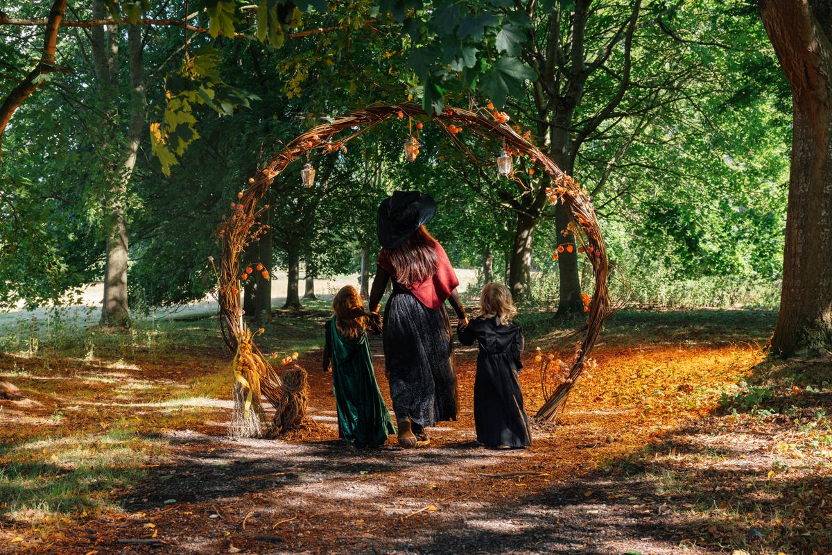 A woman and 2 children all dress up as witches walking through a wooden archway in a woodland