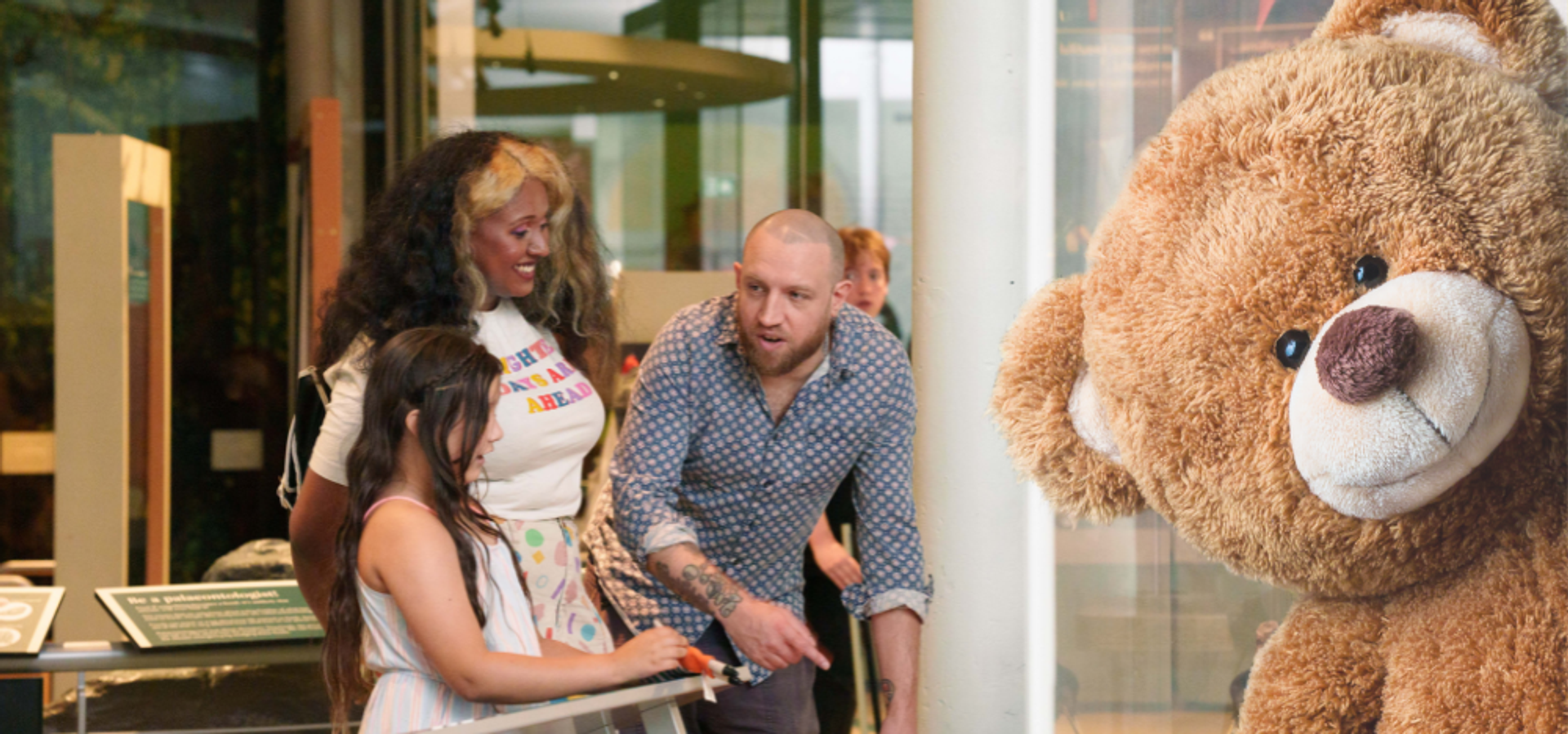 Two adults and a child looking at a museum display with a large teddy bear peaking in