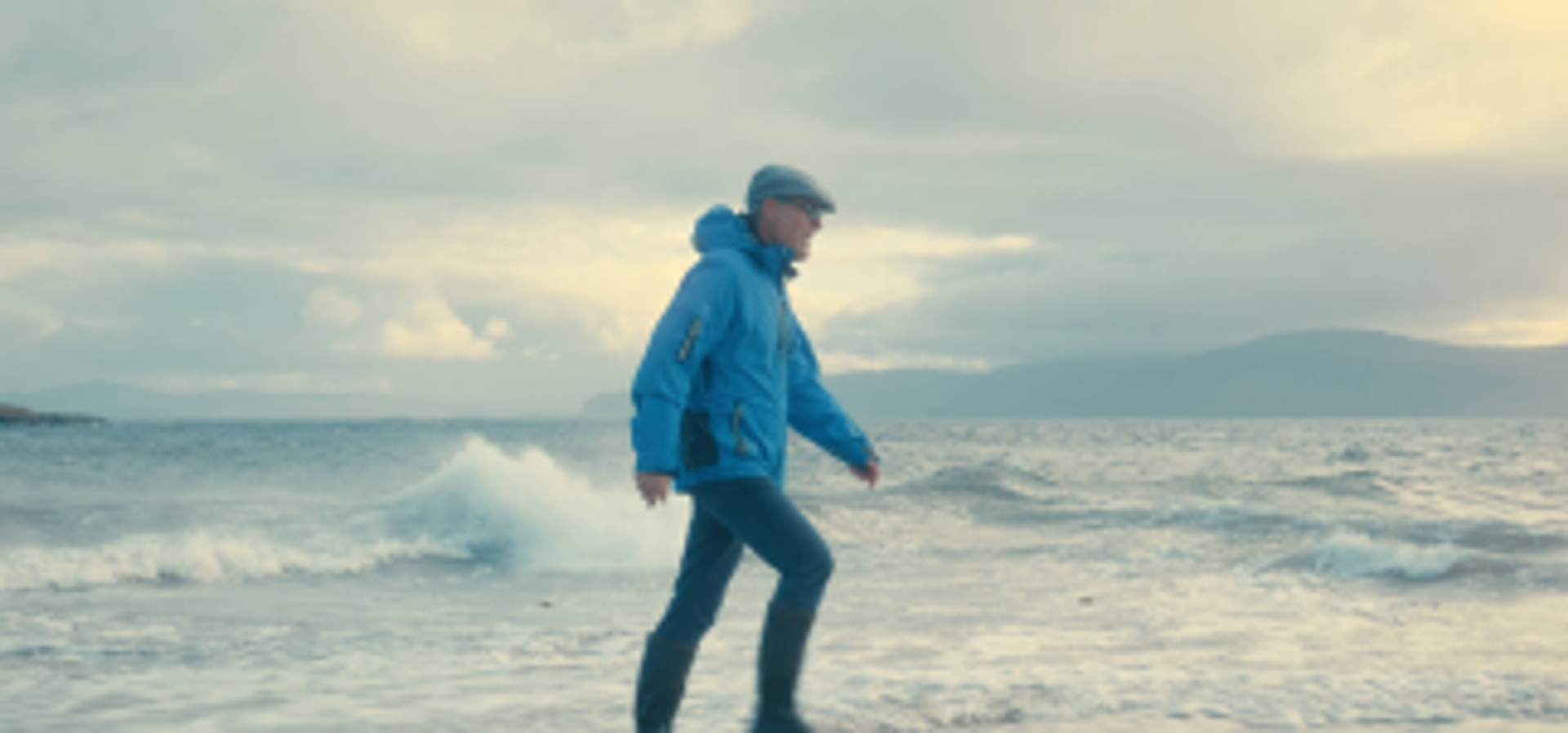 Photograph of male figure walking, with a background of a rough sea and cloud sky.