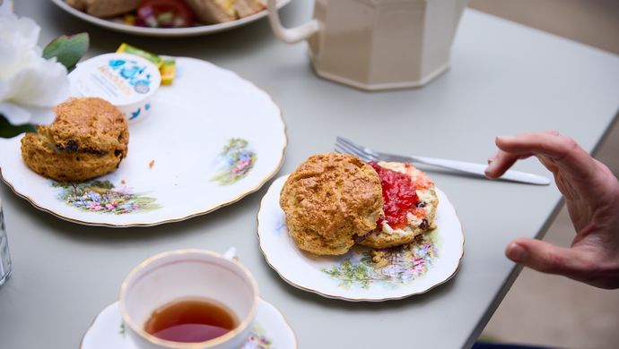 a hand reaching for a scone covered in jam and cream, with a cup of tea on the table an another plate with an extra scone on