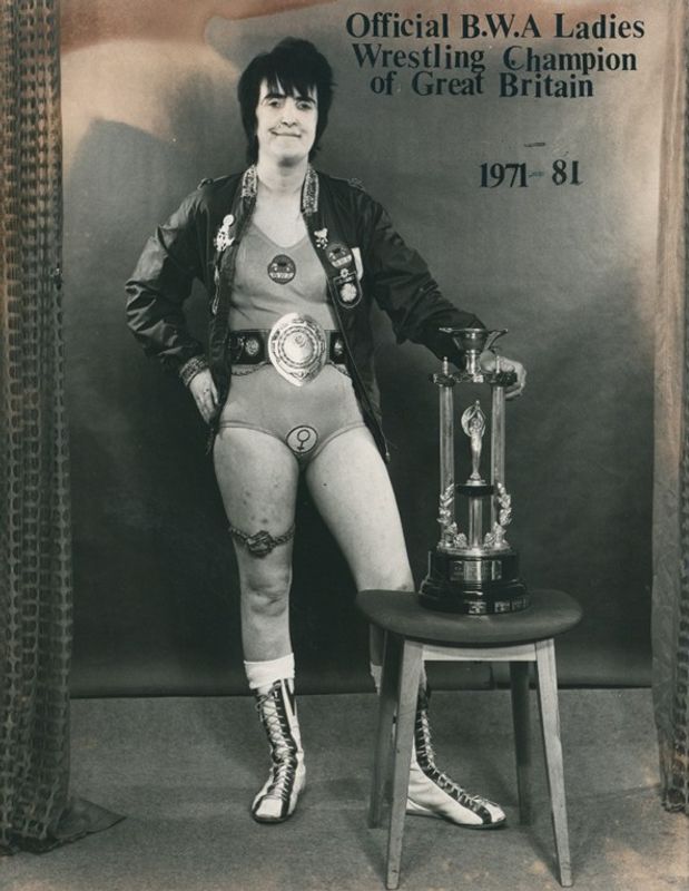 Sue Brittain stood with a trophy in front of a wall that says 'Official B.W.A Ladies Wrestling Champion of Great Britain'