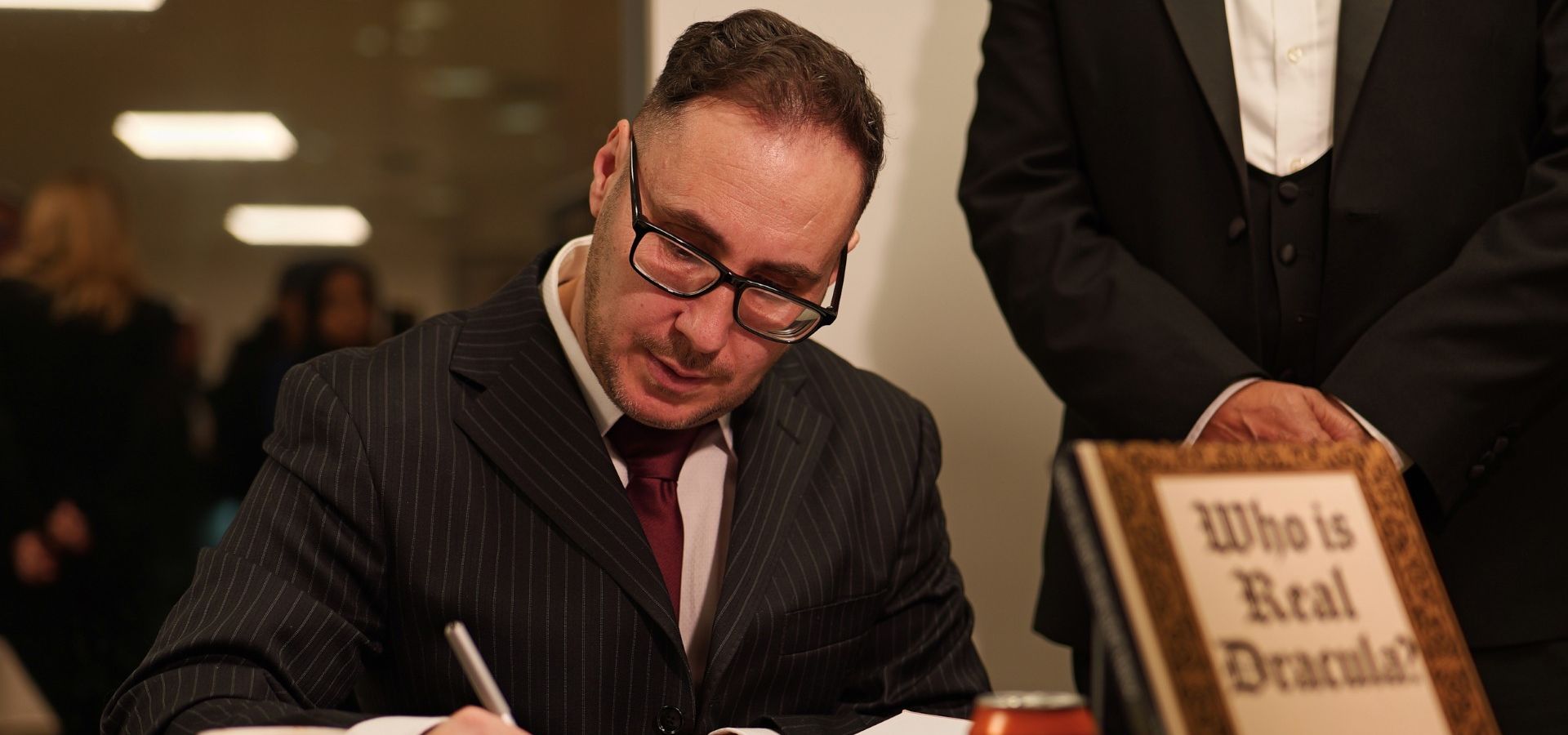 A man writing in a book with a sign in front saying who is real dracula?
