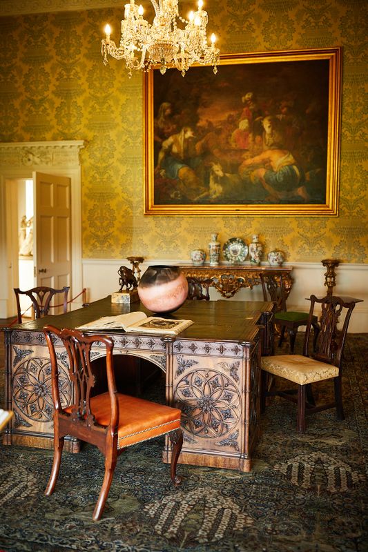 A wallpapered room with a large 4 person desk, decorative items and a painting on the wall