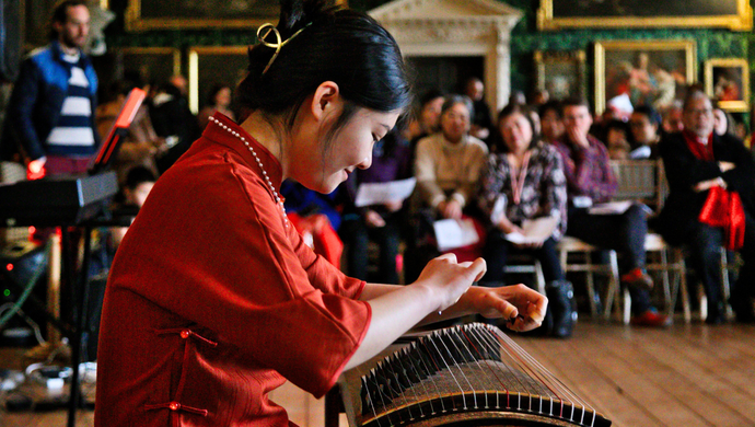 A woman banging on a drum in a red top, surrounded by people sat down in Temple Newsam