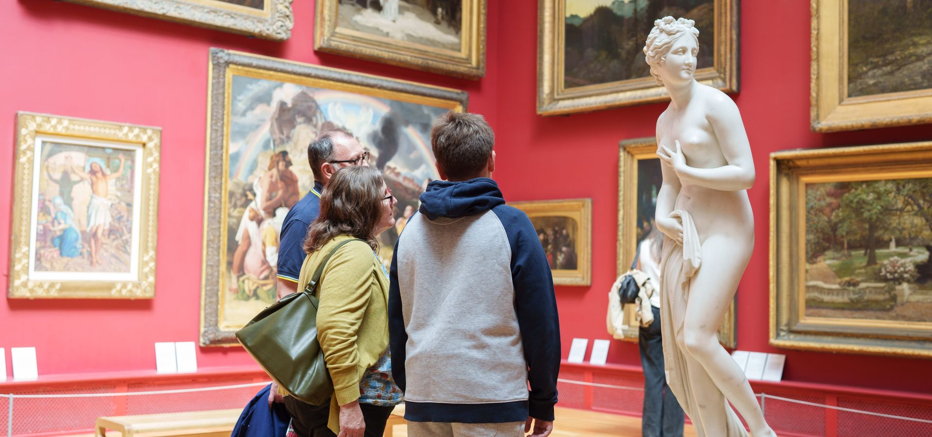 3 people looking at a sculpture of a woman in a red room with framed paintings on the wall