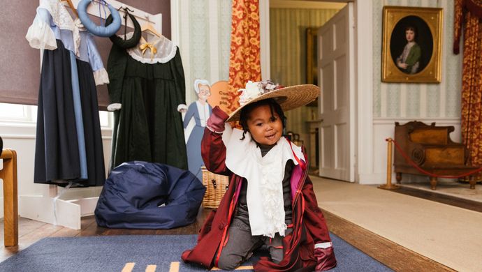 A toddler kneeling on the floor in fancy dress wearing a hat